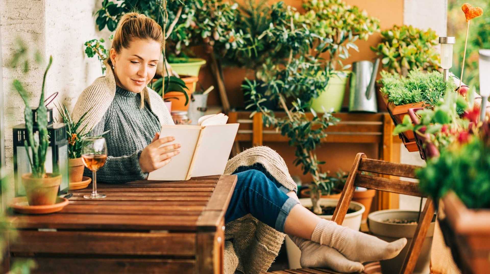 Natürliche Holzgartenmöbel betten sich in die Gartenlandschaft ein. Eine Frau genießt ihr Buch und ihren Cocktail am Gartentisch.