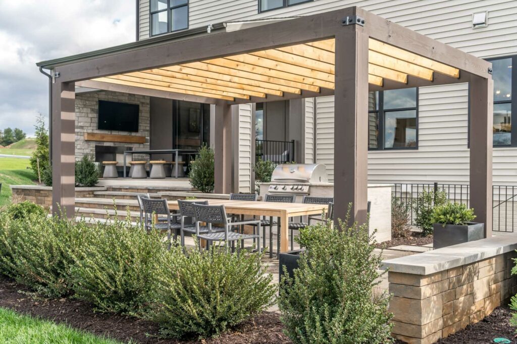 Terrassenüberdachung aus Holz und Metall im Garten. Darunter befindet sich eine gemütliche Sitzecke mit Grill.