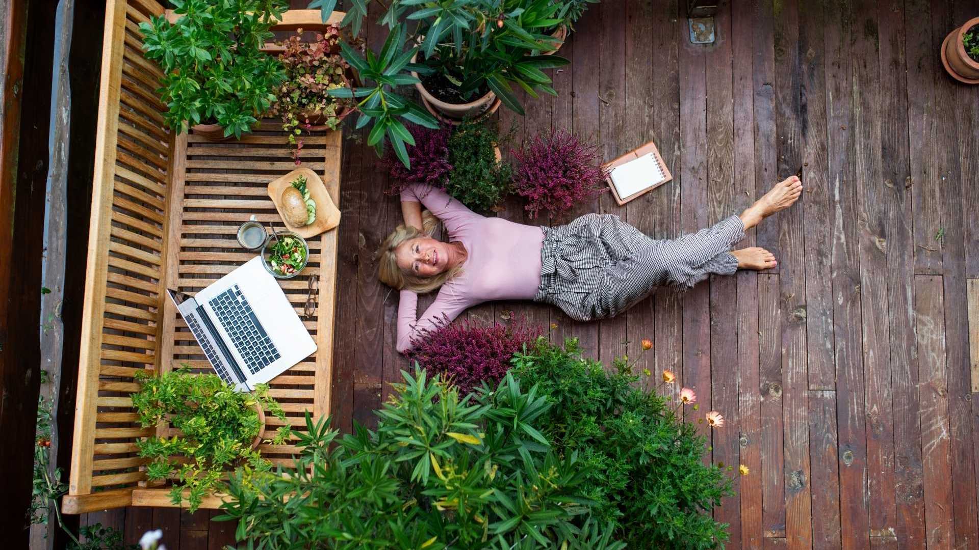 Seniorin entspannt auf eigener Terrasse mit Essen, Laptop und wirkt gelassen inmitten von Pflanzen.